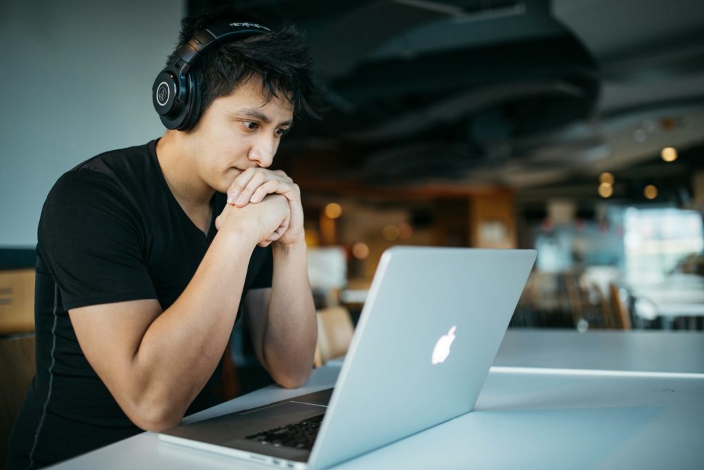 Junger Mann mit Kopfhörern und schwarem T-Shirt sitzt vor einem MacBook und hat dabei den Kopf auf seinen Händen abgestützt, als ob er etwas lesen oder ein Video schaut.