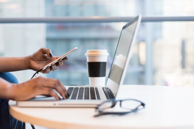 Fraunenhände einer schwarzhäutigen Frau, die in der linken Hand auf einem Smartphone tipp und mit die rechte Hand auf der Tastatur eines Laptops hält. Davor liegt eine Lesebrille und im Hintergrund ist ein Coffe-to-Go-Becher zu sehen