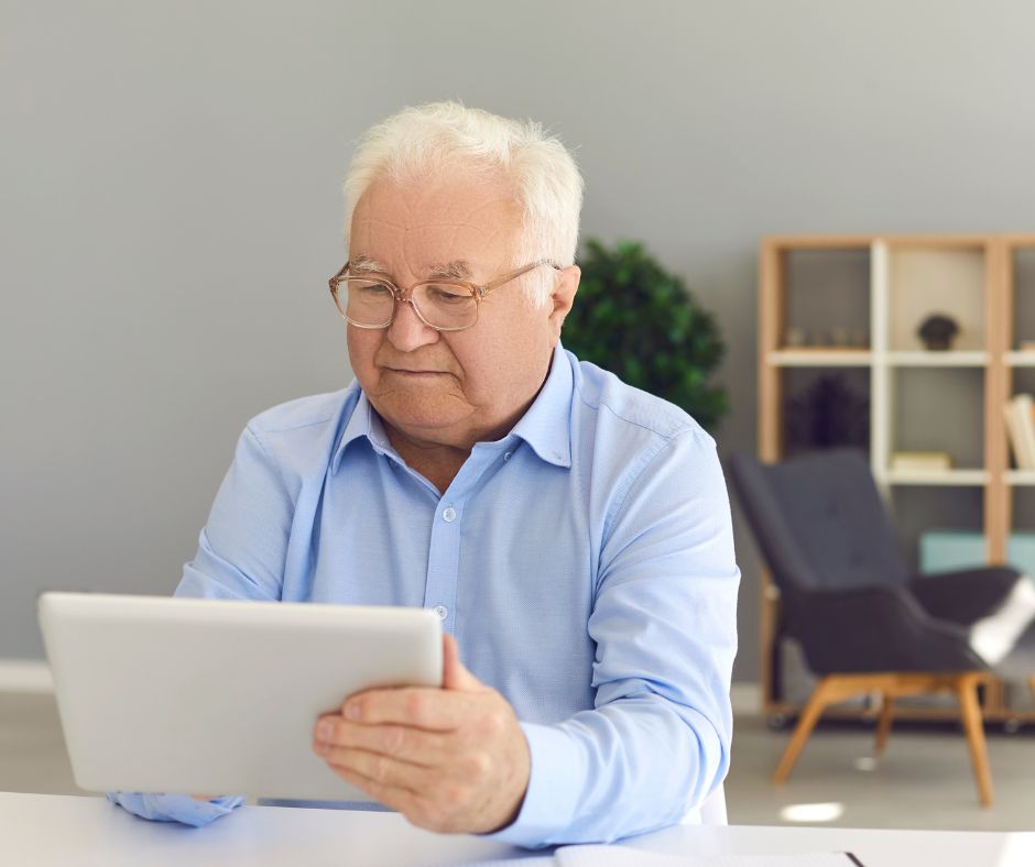 Ein alter Herr sitzt am Schreibtisch zu Hause und hält ein Tablet in der Hand und nutzt das Internet, z. B. um einen Termin online beim Arzt zu vereinbaren.