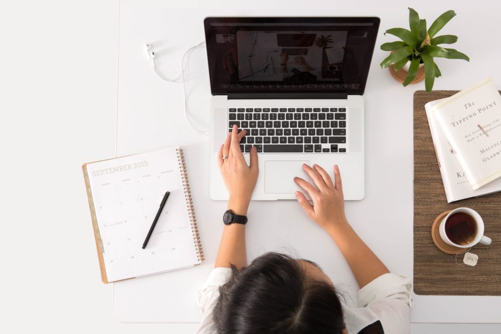 Eine Frau sitzt am Schreibtisch, vor ihr sehet ein aufgeklappter Laptop. Daneben ein Notizbuch, eine Pflanze und Kaffe
