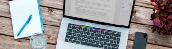 Aufgeklappter Laptop auf einem Holztisch, daneben liegt ein Notizblock sowie ein Smartphone. Außerdem steht ein Wasserglas und eine Blume auf dem Tisch. Auf dem Screen des Laptops sieht man einen Text.
